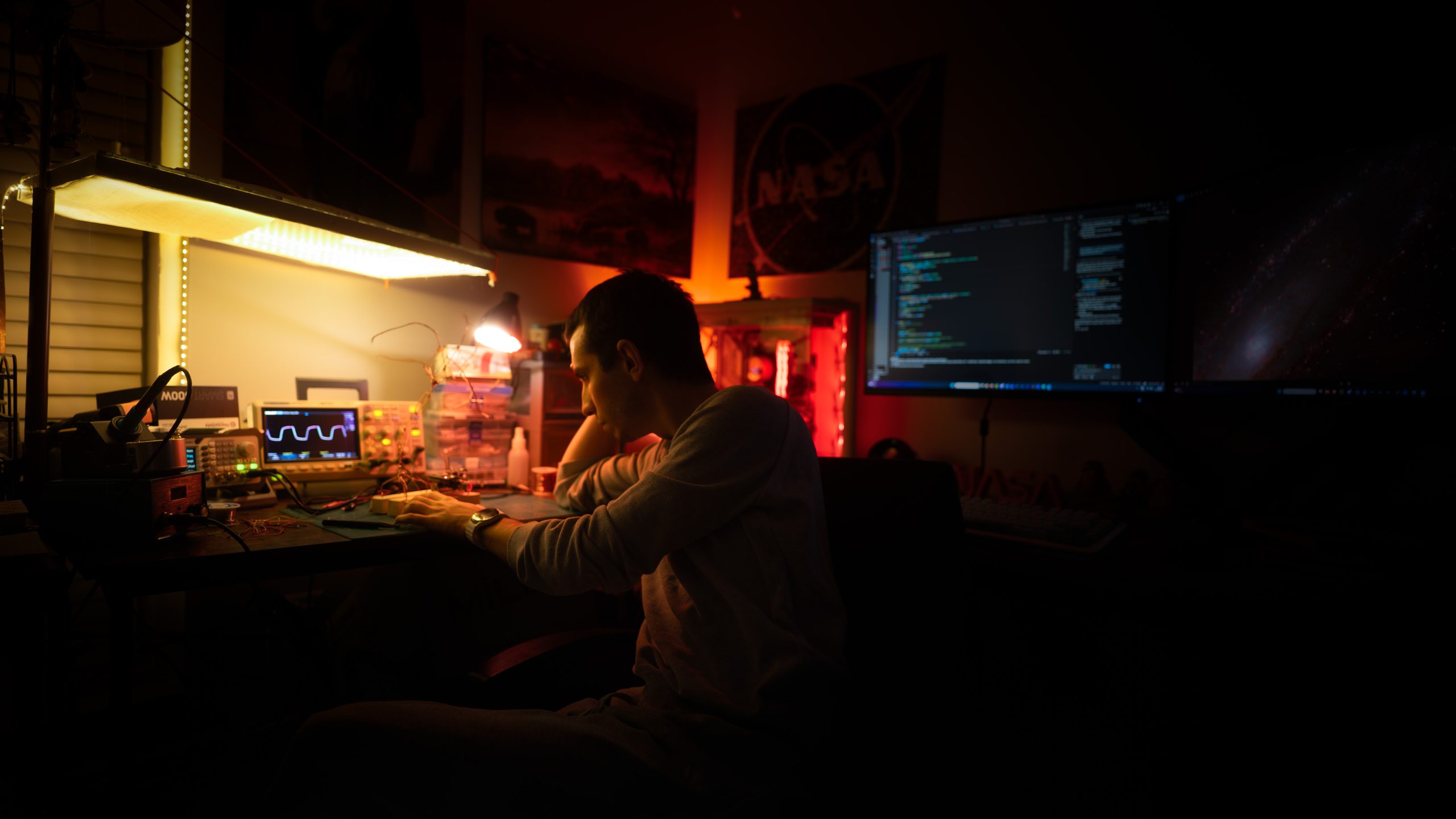 Ogetay Kayali at a workbench, with an oscilloscope, a desk lamp, and a NASA poster